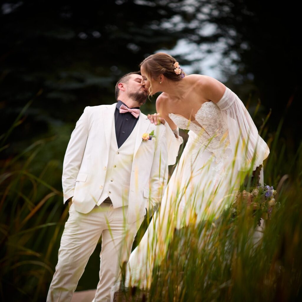 Wedding couple sharing a kiss framed through tall reeds by the pond, Lazy Stud Ranch Lyons Colorado